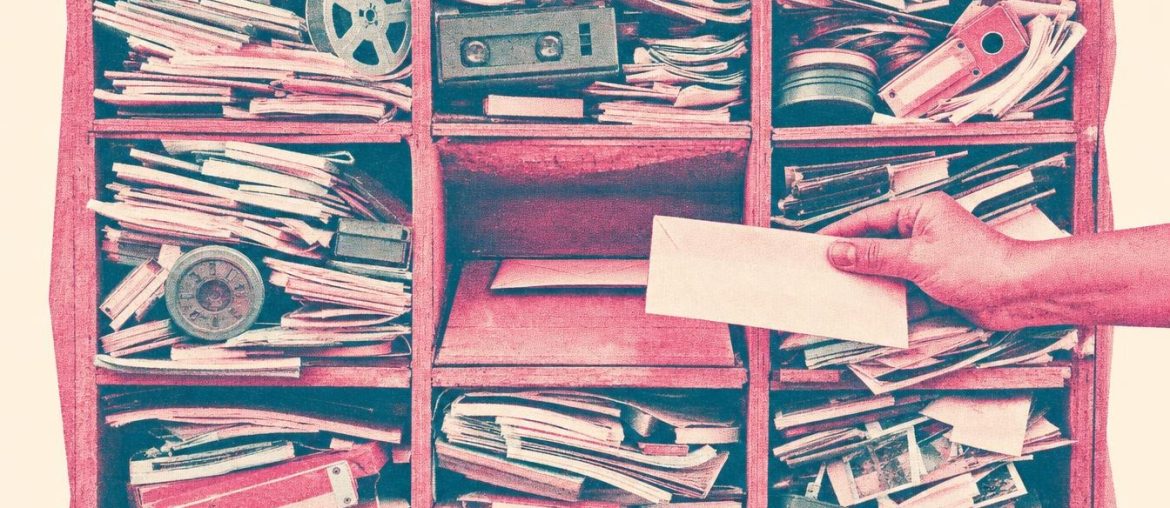 Sorting shelf with eight cubbies overflowing with photos, film reels and binders, and a nearly empty middle cubby where a hand slips in a letter